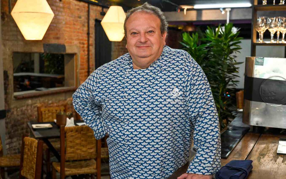 O chef Erick Jacquin com mão na cintura, uniforme de chef de cozinha, olhando com leve sorriso para a câmera