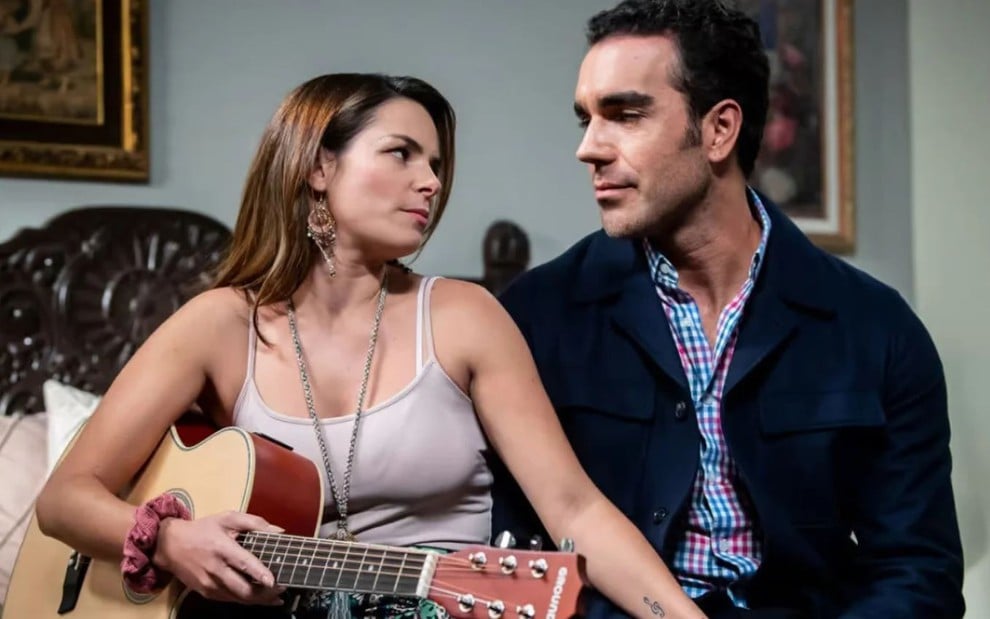 Os atores Alejandra Robles Gil e Marcus Ornellas na novela Eternamente Apaixonados, se olhando, sérios, ela com violão no colo