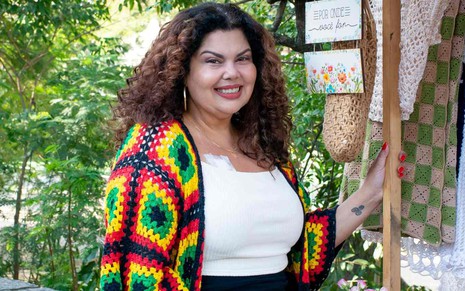 A atriz Fabiana Karla sorrindo, com casaco colorido de crochê, em frente a barraca de camelô, em cena do filme Câncer com Ascendente em Virgem