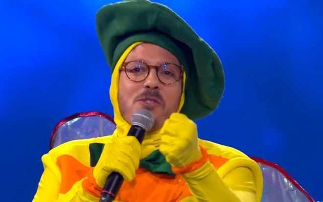 Fabio Porchat fala em microfone; ele está vestido com traje amarelo e laranja do personagem Dengue