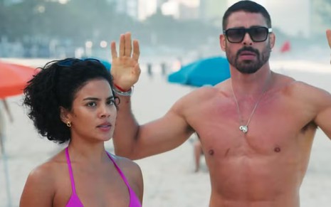 A atriz Bella Campos séria e o ator Cauã Reymond ao lado dela, com mãos ao alto, em cena de Vale Tudo
