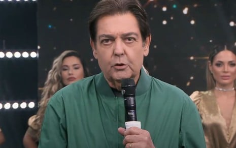 Fausto Silva no cenário do Faustão na Band, com casaco verde de gola alta e segurando microfone