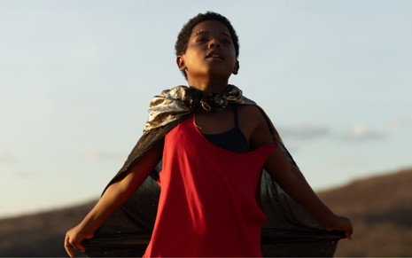 menino negro abre os braços em paisagem natural; ele olha para o céu; ele veste uma camiseta vermelha e uma capa