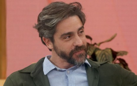 Felipe Abib sentado no sofá do Encontro, usando casaco verde-escuro e camisa azul-claro