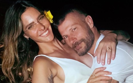 Fernanda Lima e Rodrigo Hilbert abraçados e sorridentes na véspera de Ano-Novo; os dois vestem branco e ela usa uma flor amarela no cabelo