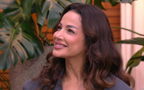 mulher branca sorri em estúdio de TV; ela veste uma blusa cinza