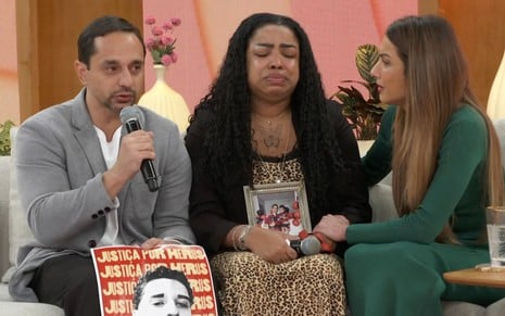 Fernando Guimarães e Mônica Guimarães sentados no sofá do Encontro, enquanto Patrícia Poeta segura a mão da mulher, que chora, e o homem segura um pôster do filho Herus