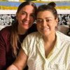 Flora Gil e Preta Gil juntas, sentadas em um sofá, dando as mãos e sorrindo