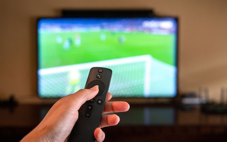 Mão segura controle remoto diante de TV que exibe jogo de futebol com imagem desfocada