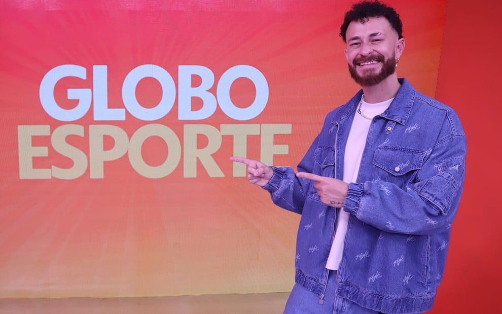 Fred Bruno usa jaqueta e calça jeans e uma camiseta branca; ele encara a câmera, sorrindo, enquanto aponta para o logo do Globo Esporte