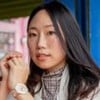Gabi Yoon sentada em cenário de Volta por Cima; ela usa blazer branco, blusa xadrez e relógio claro no pulso, além de estar com as mãos próximas ao rosto