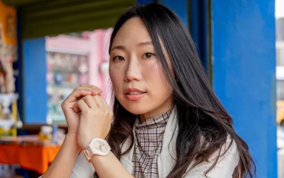 Gabi Yoon sentada em cenário de Volta por Cima; ela usa blazer branco, blusa xadrez e relógio claro no pulso, além de estar com as mãos próximas ao rosto