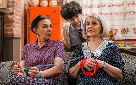 Felipe (Gabriel Avellar) está atrás do sofá e fala com Shirley  (Rosi Campos) e Wanda (Magali Biff) em cena da novela A Caverna Encantada