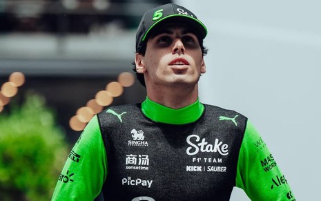 Homem de boné preto e macacão verde com preto aparece em pista de corrida; ele olha para cima com expressão séria.