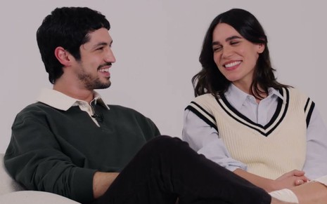 Gabriel Leone e Carla Salle sentados em um fundo branco, olhando um para o outro e rindo