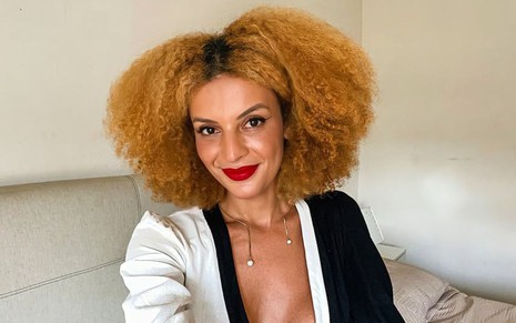 Gabriela Loran está com cabelos ruivos soltos e sorri para a foto; ela usa um vestido preto e branco e batom vermelho