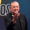 Galvão Bueno no estúdio de Galvão e Amigos; ele aparece todo de preto, estendendo a mão direita, em frente à logo do programa