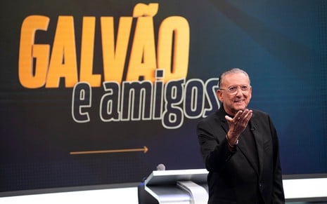 Galvão Bueno no estúdio de Galvão e Amigos; ele aparece todo de preto, estendendo a mão direita, em frente à logo do programa