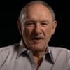 Gene Hackman em entrevista ao YouTube; ator usa camisa cinza e está em cenário totalmente preto