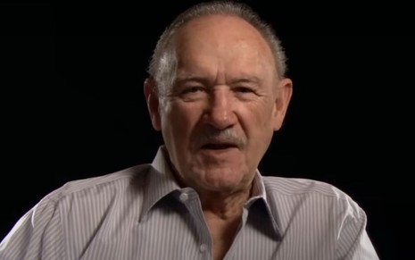 Gene Hackman em entrevista ao YouTube; ator usa camisa cinza e está em cenário totalmente preto