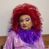Homem vestido de drag queen usa peruca vermelha, maquiagem pesada e roupa brilhante com plumas roxas, posando para duas fotos.