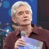 Homem de cabelo grisalho segura microfone e plaqueta do programa "Altas Horas" em estúdio com plateia e banda ao fundo.