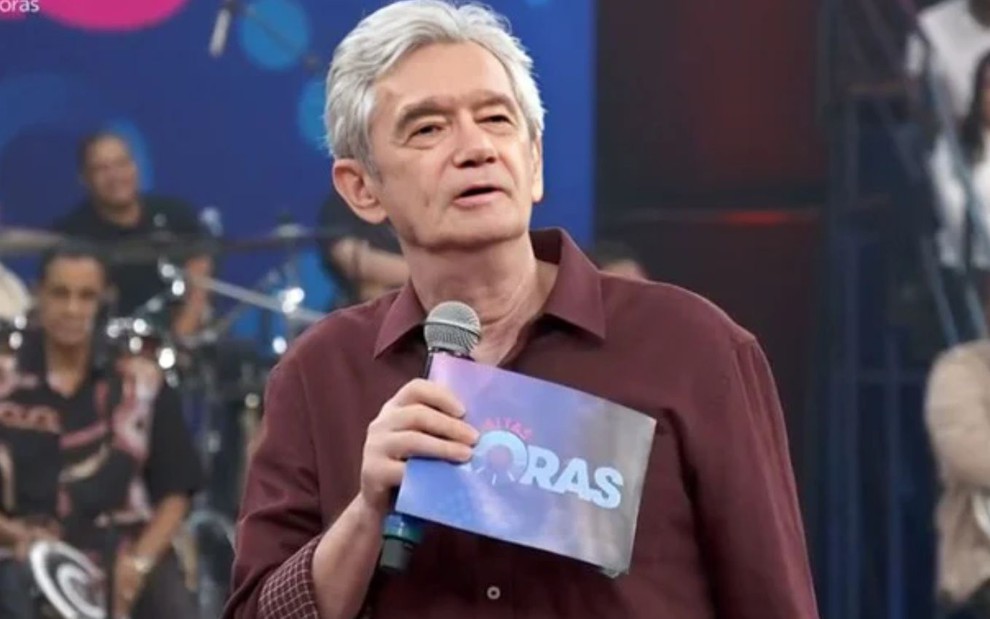 Homem de cabelo grisalho segura microfone e plaqueta do programa "Altas Horas" em estúdio com plateia e banda ao fundo.