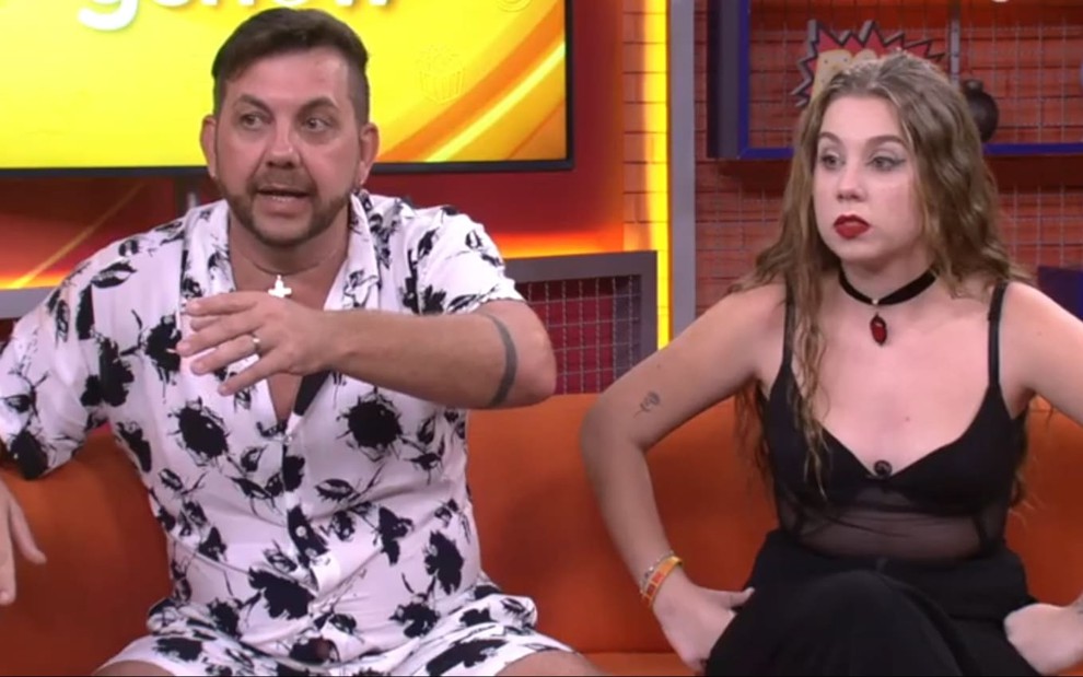 Edilberto Simões usa uma camisa estampada e está sentado ao lado da filha, Raissa Simões, que usa um body preto de alcinha; os dois exprimem choque