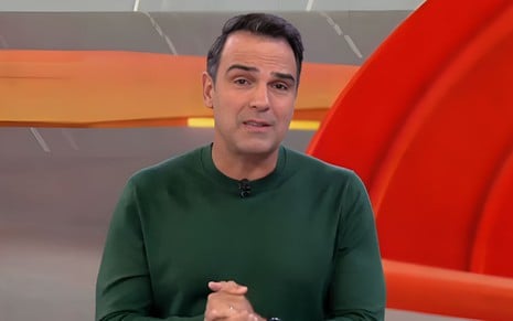 Homem com cabelo curto, usando blusa verde escura e microfone preso na gola, em ambiente com tons laranja e cinza ao fundo.