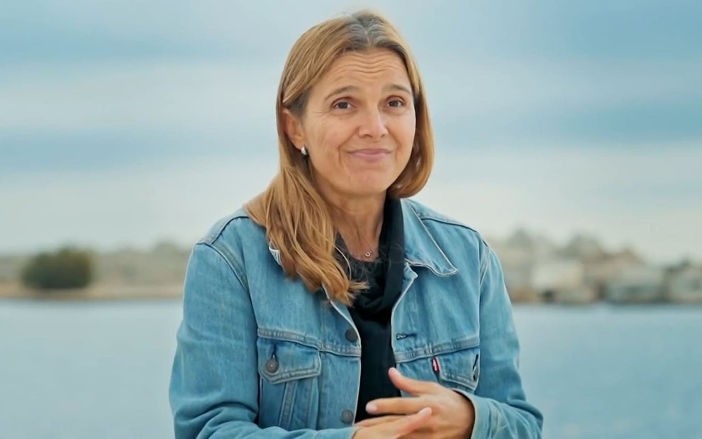 Vanessa Cristina Soares usa uma camiseta preta e uma jaqueta jeans; ela está na frente de uma praia em Barcelona, Espanha