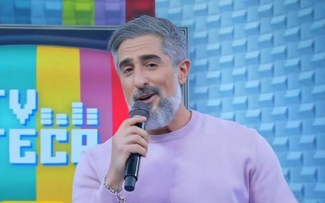 Homem de cabelo grisalho segura microfone e sorri em estúdio colorido com fundo azul e letreiro “TV TECA” ao fundo.