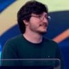 Homem de barba e óculos redondos sorri de leve em um estúdio de TV com fundo colorido. Ele veste suéter verde-escuro.        Perguntar ao ChatGPT