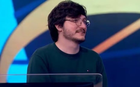 Homem de barba e óculos redondos sorri de leve em um estúdio de TV com fundo colorido. Ele veste suéter verde-escuro.        Perguntar ao ChatGPT