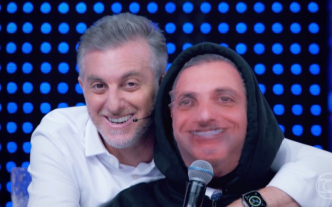 Luciano Huck abraça Ed Gama, que está usando uma máscara com o rosto do apresentador