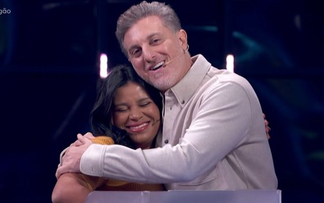 Luciano Huck abraça a participante Eliza no palco do Domingão; os dois sorriem emocionados, com expressão de carinho e apoio.