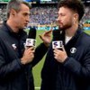 Edgar Alencar conversa com Fred Bruno em frente a um campo de futebol; eles estão com casacos pretos e microfones nas mãos