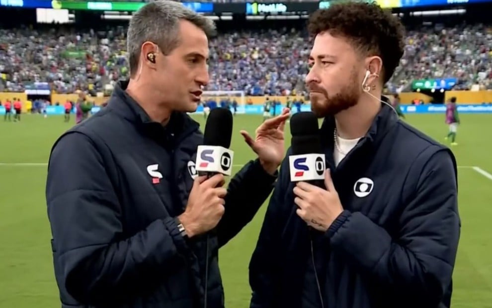 Edgar Alencar conversa com Fred Bruno em frente a um campo de futebol; eles estão com casacos pretos e microfones nas mãos