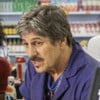 Em um cenário de supermercado, vários atores estão reunidos em torno de um caixa, com expressões de interesse
