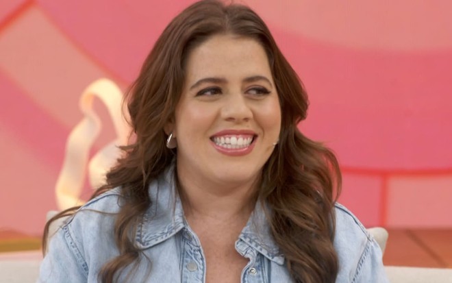 Tati Machado usa uma blusa vermelha e uma jaqueta jeans clara; ela está sorridente