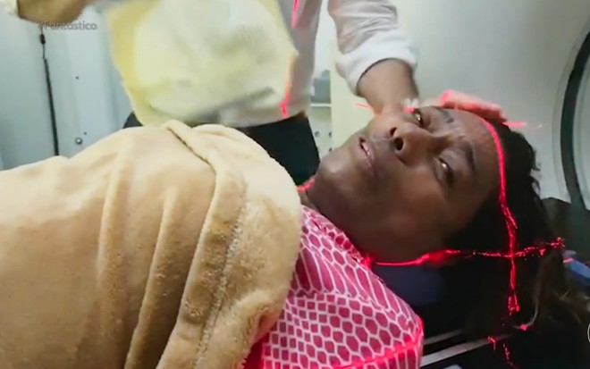 Gloria Maria está deitada em uma câmara de ressonância, com raios lasers vermelhos formando um quadrado em sua cabeça