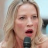 Mulher loira, de olhos claros e expressão surpresa, segura um microfone preto. Ela está em ambiente claro, com fundo desfocado.