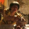 Senhora força homem imobilizado a comer pão; cena ocorre em ambiente retrô com luz baixa e decoração religiosa.