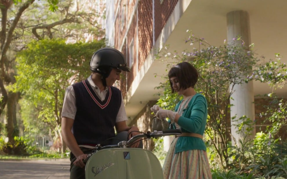 Cena de duas pessoas ao ar livre, ao lado de uma moto Vespa. Ele usa capacete e suéter; ela usa cardigã e parece mexer em uma bolsa.