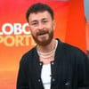 Homem em frente ao fundo do programa "Globo Esporte", vestindo um casaco preto com símbolo da Nike, colar de pérolas e barba cheia.