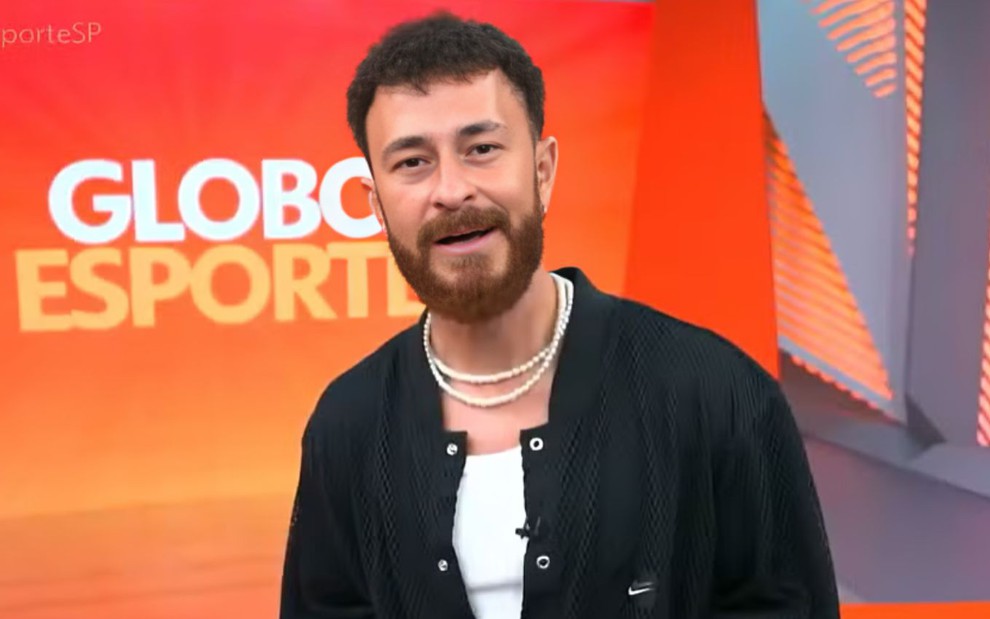 Homem em frente ao fundo do programa "Globo Esporte", vestindo um casaco preto com símbolo da Nike, colar de pérolas e barba cheia.