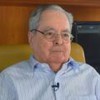 Um senhor de idade, com óculos e cabelos brancos, está sentado em uma poltrona em uma sala