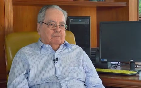 Um senhor de idade, com óculos e cabelos brancos, está sentado em uma poltrona em uma sala
