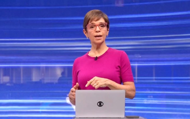 Apresentadora em estúdio de TV, com fundo azul em linhas, veste blusa rosa e óculos. Está em frente a um laptop com logotipo