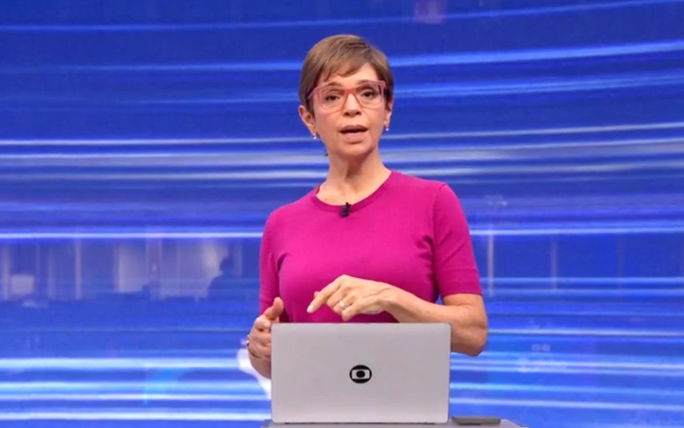 Apresentadora em estúdio de TV, com fundo azul em linhas, veste blusa rosa e óculos. Está em frente a um laptop com logotipo