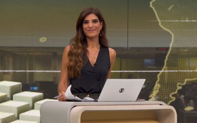 Andreia Sadi no cenário do Jornal Hoje, sorri, com um computador aberto à sua frente